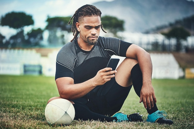 Há tempo para o rugby, depois há tempo para conectividade Foto de corpo inteiro de um jovem e bonito jogador de rugby usando um smartphone enquanto está sentado no campo de jogo durante o dia