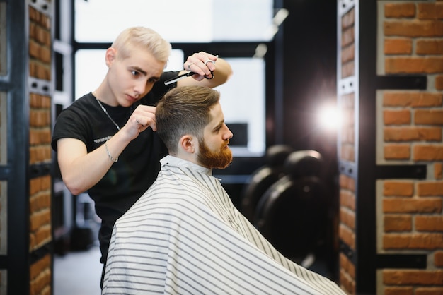 Haarschnitt perfekt aussehen lassen. Junger bärtiger Mann, der Haarschnitt durch Friseur erhält, während im Stuhl am Friseursalon sitzt