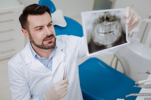 Foto hábil y experimentado dentista prominente que estudia la imagen de rayos x de los dientes de sus pacientes mientras averigua el diagnóstico antes de comenzar un tratamiento