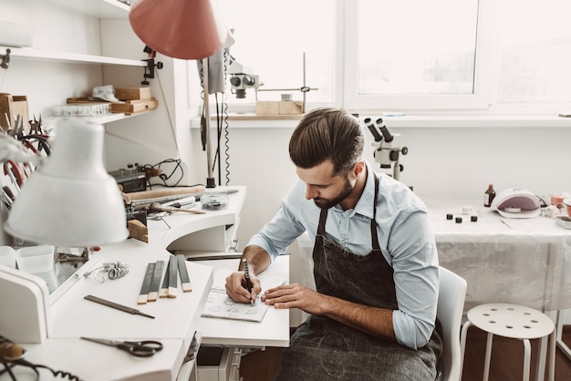 Habilidades artísticas. Joven joyero barbudo está dibujando un boceto en su taller