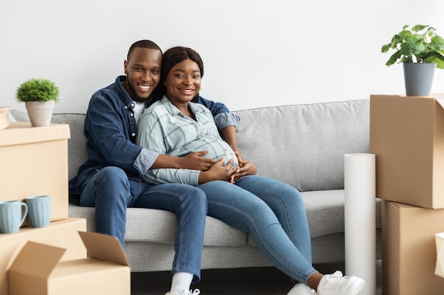 Habitação familiar. cônjuges grávidas negras felizes posando no sofá em novo apartamento