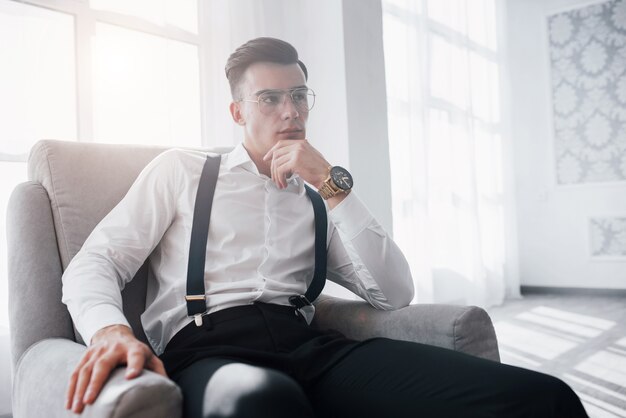 Habitacion amplia. Bonito reloj en la mano. Cerrar foto de chico atractivo en las gafas y ropa elegante sentado en la silla.