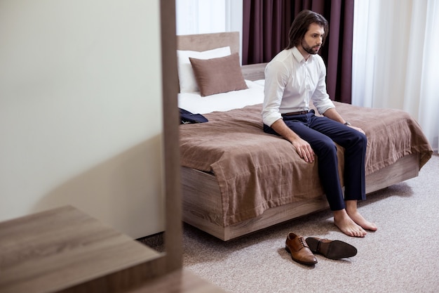 En la habitación del hotel. Triste hombre cansado sentado en la cama en el hotel mientras está muy cansado