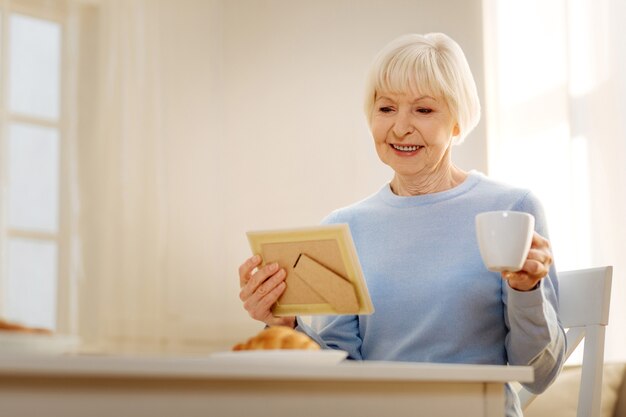 Habitación luminosa. Increíble pensionista manteniendo una sonrisa en su rostro y mirando la foto mientras está sentado en el comedor