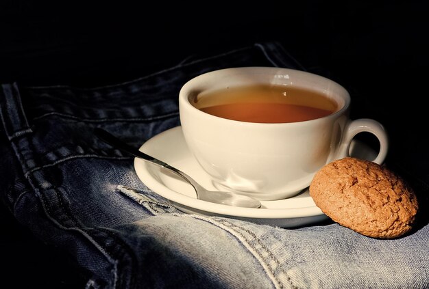 Hábitos saudáveis Conceito de hora do chá Caneca de xícara de água quente e saco de chá Caneca cheia de água fervente e saquinho de chá no fundo de jeans azul Folha inteira verde ou preta de ervas Processar a fabricação de chá em caneca de cerâmica