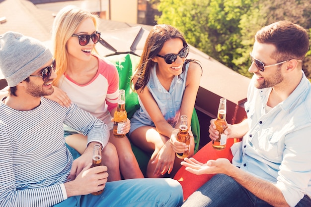 Habla sin preocupaciones con tus amigos. Vista superior de cuatro jóvenes alegres charlando y bebiendo cerveza mientras están sentados en las bolsas de frijoles en el techo del edificio