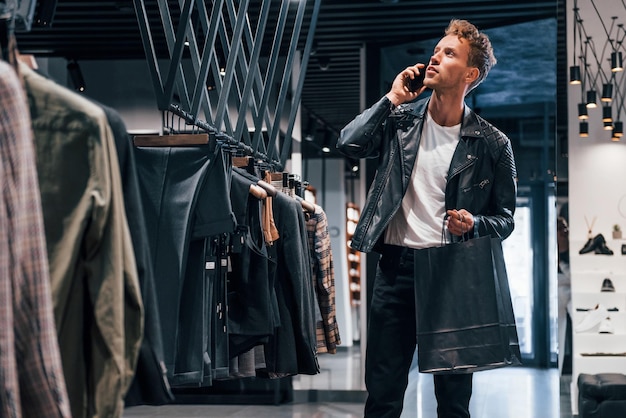 Habla por teléfono Chico joven en una tienda moderna con ropa nueva Ropa elegante y cara para hombres