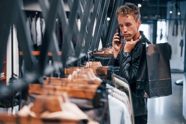 Habla por teléfono Chico joven en una tienda moderna con ropa nueva Ropa elegante y cara para hombres