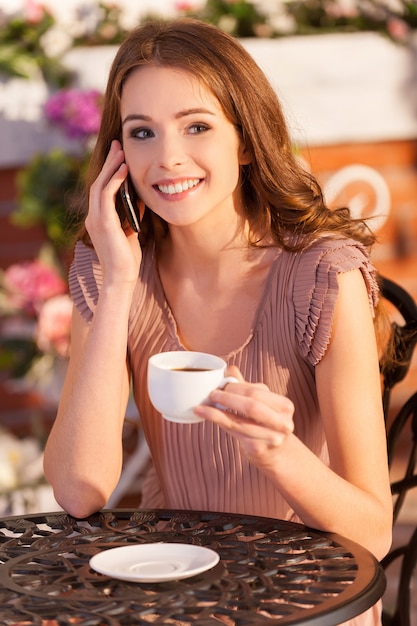 Hablando con un amigo. Atractiva mujer joven hablando por teléfono móvil y sonriendo mientras está sentado en el café al aire libre