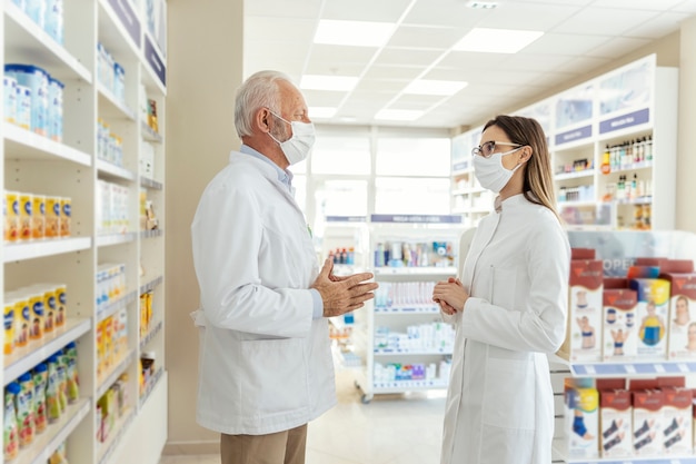 Hablando entre dos farmacéuticos y el virus corona. Antes de jubilarse, el farmacéutico le enseña al joven farmacéutico cómo trabajar en una farmacia. Llevan uniformes y mascarillas