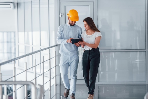 Hablando con una joven con ropa informal Ingeniero con ropa blanca y casco protector naranja de pie y trabajando en interiores