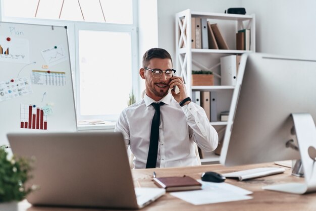 Hablar con el cliente. Apuesto joven en ropa formal hablando por teléfono y sonriendo mientras está sentado en la oficina