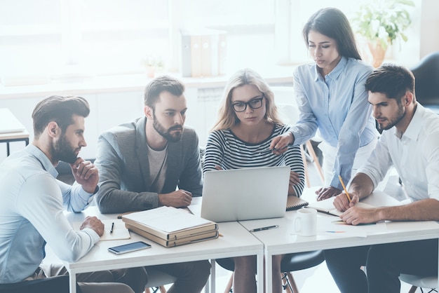 Hablar de cuestiones comerciales. Grupo de jóvenes empresarios sentados en el escritorio de la oficina y discutiendo algo mientras miran juntos la computadora portátil