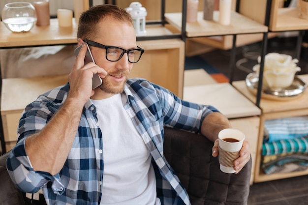 Hablar con la gente. Hombre positivo barbudo guapo sosteniendo un vaso de plástico y tomando café mientras habla por teléfono