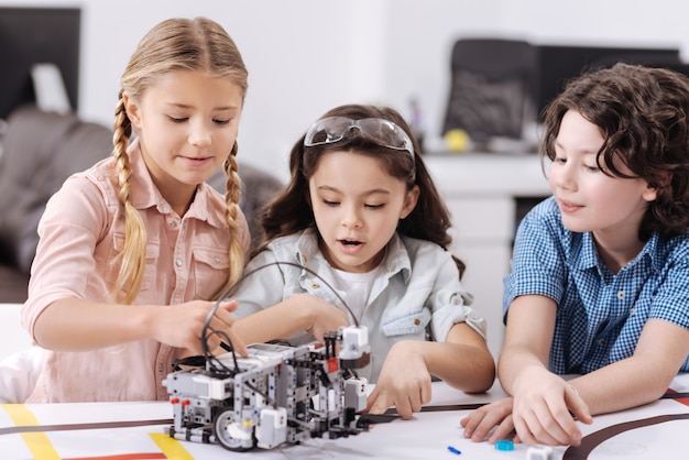 Hablar de investigación tecnológica. Pequeños científicos activos alegres sentados en la escuela y disfrutando de la clase de tecnología mientras construyen un robot