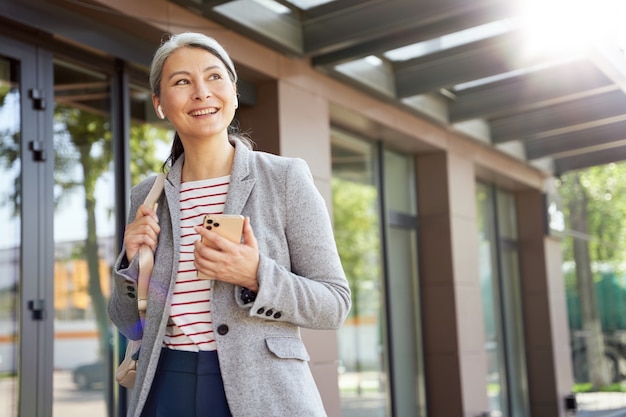 Hablar de negocios. Retrato de una mujer hermosa y feliz con auriculares inalámbricos con smartphone, hablando con alguien por teléfono mientras está de pie al aire libre. Gente de negocios, trabajo