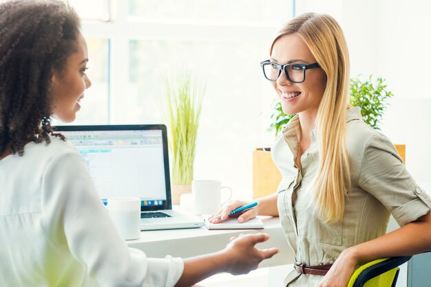 Hablar sobre el trabajo en equipo. Dos mujeres jóvenes felices discutiendo algo mientras están sentados juntos en su lugar de trabajo