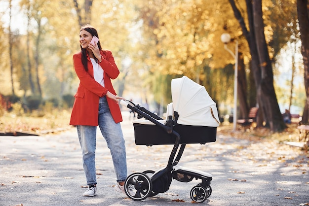 Hablar por teléfono. Madre con abrigo rojo pasea con su hijo en el cochecito en el parque en otoño.