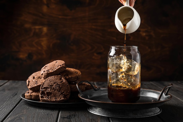 Hacer café helado. Verter café negro de una jarra de leche en un vaso. Galletas de chocolate en un plato.