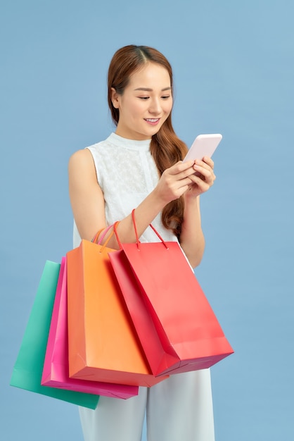 Hacer la conexión con el comercio minorista. Hermosa mujer joven con bolsas de la compra usando su teléfono inteligente con una sonrisa mientras está de pie