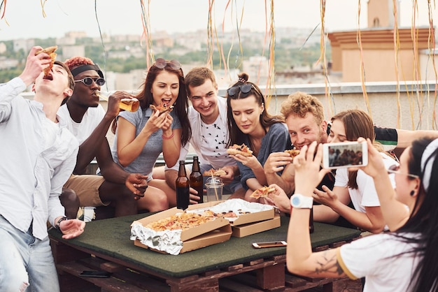 Hacer una foto Pizza deliciosa Un grupo de jóvenes con ropa informal tienen una fiesta en la azotea juntos durante el día