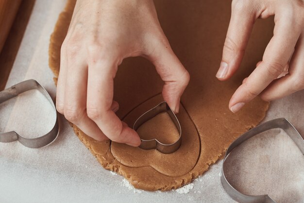 Hacer galletas de jengibre en forma de corazón para el día de San Valentín. Cortador de galletas de uso de mano de mujer. Concepto de comida de vacaciones