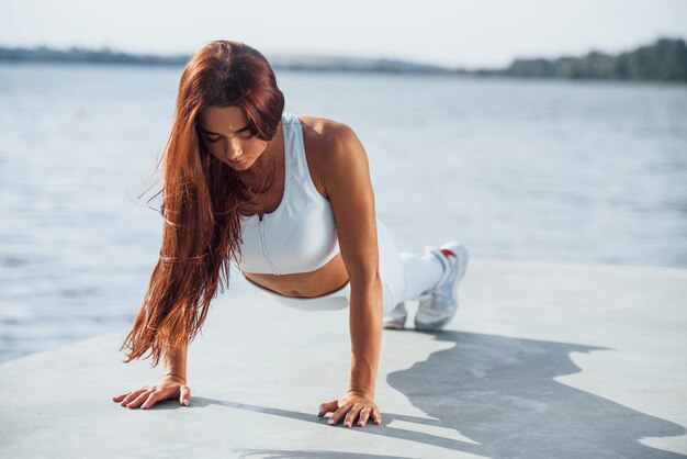 Haciendo flexiones. Foto de mujer deportiva haciendo ejercicios de fitness cerca del lago durante el día.