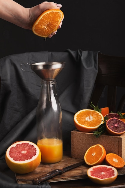 Haciendo jugo de naranja, naturaleza muerta. Composición con varios cítricos, tarro y cuchillo en silla rústica negra. Mano femenina preparando espacio fresco, copia