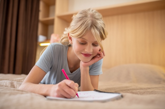 Haciendo lecciones. Una niña acostada en la cama y escribiendo algo en un cuaderno