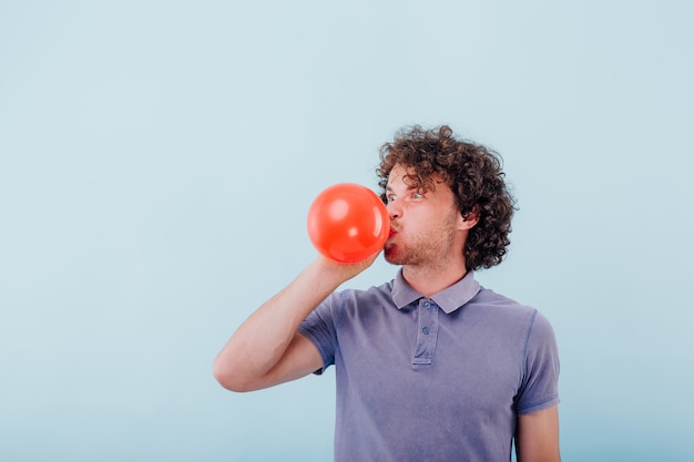 Haciendo muecas al hombre sin afeitar con el pelo rizado en camisa azul soplando globo rojo