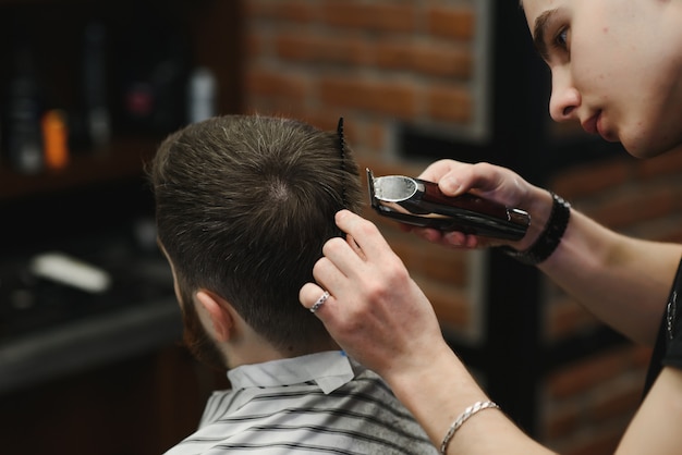 Haciendo que el corte de pelo se vea perfecto. Hombre barbudo joven cortándose el pelo por peluquero mientras está sentado en una silla en la barbería
