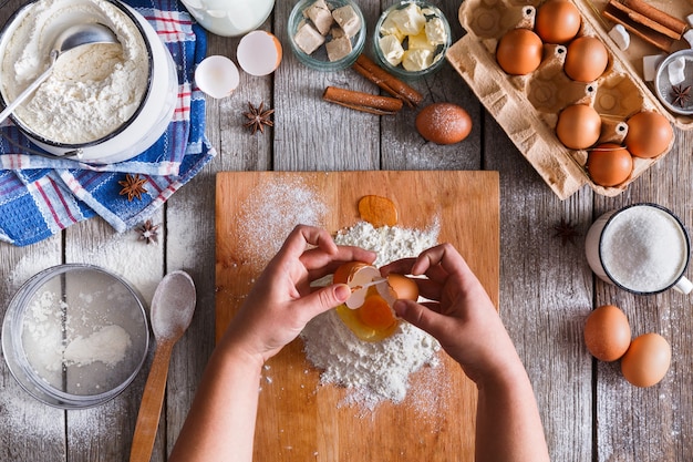 Haciendo la vista superior de la masa. Sobrecarga de las manos del panadero rompen el huevo en la harina. Cocinar ingredientes para pastelería en madera rústica, clases culinarias o concepto de receta.