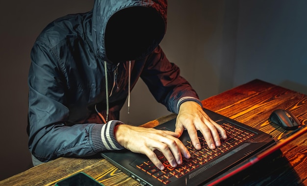 Un hacker anónimo en un capó está escribiendo en un teclado portátil en una habitación oscura El concepto de guerra cibernética y ataques Dos