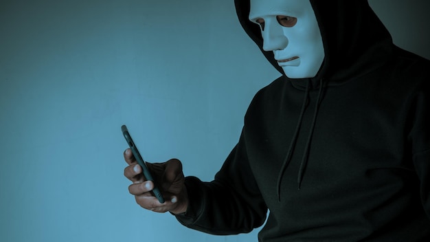 Un hacker anónimo enmascarado está usando un teléfono inteligente para penetrar la información financiera de la tarjeta de crédito Concepto de piratería y malware