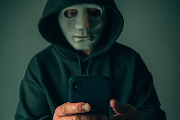 Un hacker anónimo enmascarado está usando un teléfono inteligente para penetrar la información financiera de la tarjeta de crédito Concepto de piratería y malware