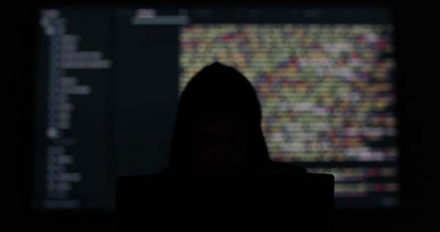 Hacker con capucha trabajando en una habitación oscura abstracta Código de programación en una pantalla grande en el fondo Seguridad del sistema digital