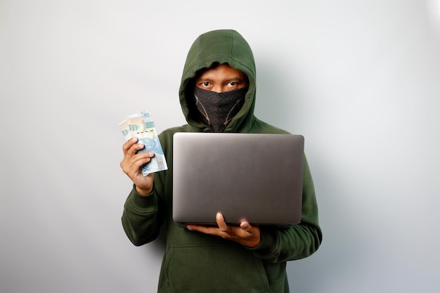 Hacker con capucha verde sosteniendo una computadora portátil y robando datos personales e información financiera mostrando dinero en la mano aislado en fondo blanco