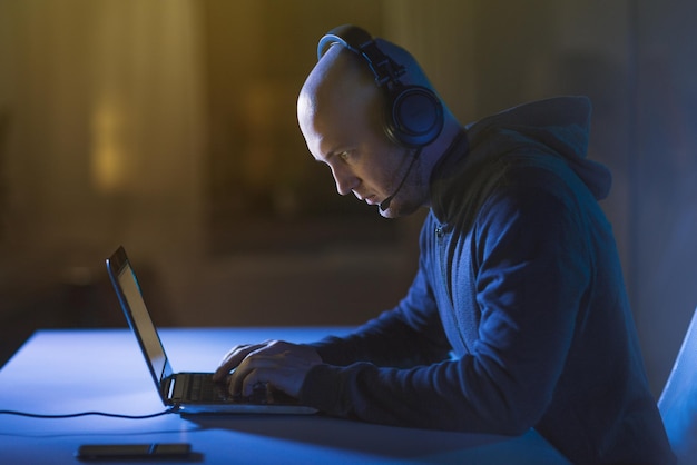 Hacker im Headset tippt auf Laptop im dunklen Raum