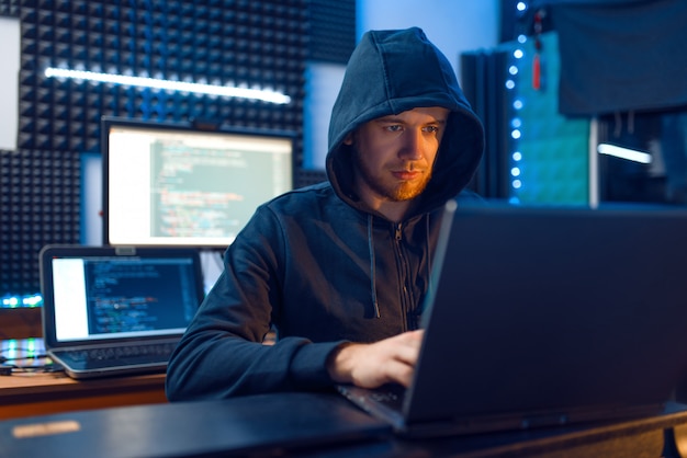 Hacker in der Haube an seinem Arbeitsplatz mit Laptop und Desktop-PC, Passwort- oder Account-Hacking, Darknet-User.