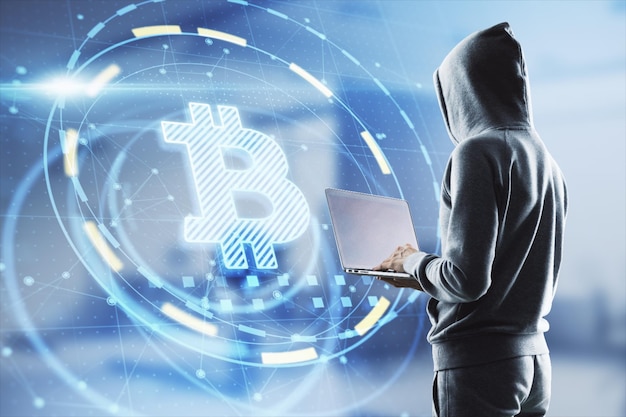 Hacker in Hoodie mit Laptop-Computer mit leuchtender Bitcoin-Schnittstelle in verschwommenem Bürointerieur Hacking-Diebstahl und Kryptowährungskonzept Doppelbelichtung