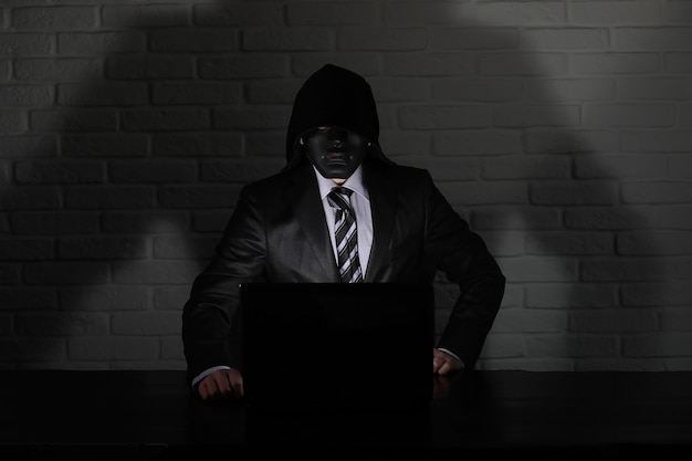 Foto hacker con máscara negra y capucha en la mesa frente al monitor