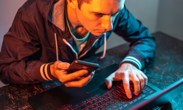 Un hacker con un teléfono está escribiendo en el teclado de una computadora portátil en una habitación oscura bajo una luz de neón