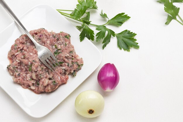 Hackfleisch und Gabel auf Teller. Geschälte Zwiebeln und Petersilie auf dem Tisch. Weißer Hintergrund. Ansicht von oben