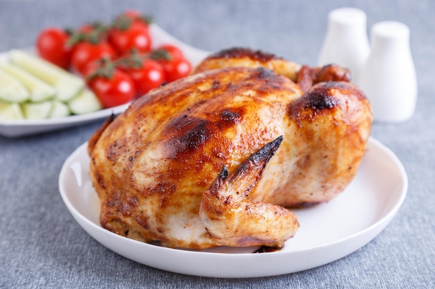 Hähnchen im Ganzen zu einer köstlichen Kruste gebacken Ein traditionelles Gericht Im Hintergrund sind Gurken und Cherry-Tomaten auf einem weißen Teller Closeup selektiven Fokus grauen Hintergrund