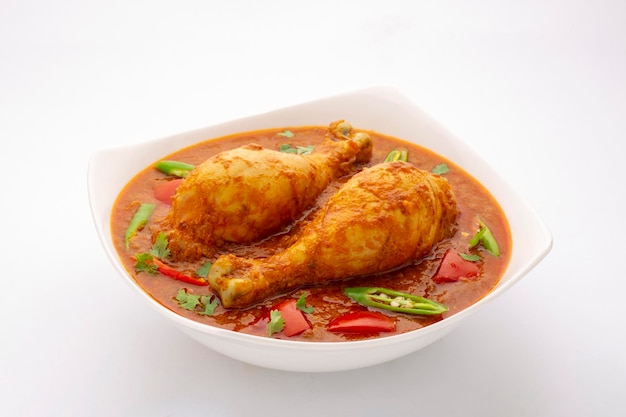 Hähnchencurry oder Masala, würziges rötliches Hähnchenschenkelgericht, garniert mit Korianderblatt und frischem grünen Chili, angeordnet in einer weißen Keramikschale mit weißem Hintergrund, isoliert.