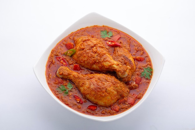 Hähnchencurry oder Masala, würziges rötliches Hähnchenschenkelgericht, garniert mit Korianderblatt und frischem grünen Chili, das in einer weißen Keramikschale mit weißem Hintergrund angeordnet ist, isoliert.