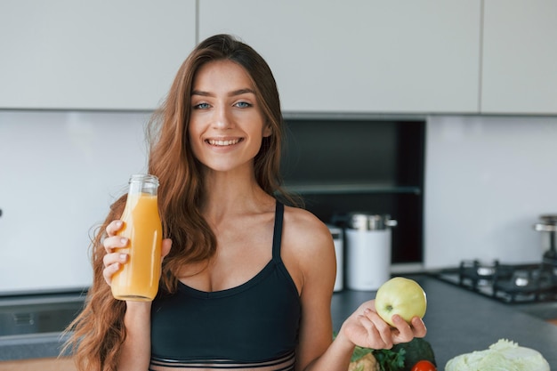 Hält Apfel in der Hand Junge Europäerin ist drinnen in der Küche drinnen mit gesunden Lebensmitteln