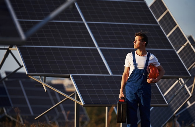 Hält Koffer mit Ausrüstung in der Hand. Männlicher Arbeiter in blauer Uniform im Freien mit Solarbatterien am sonnigen Tag.