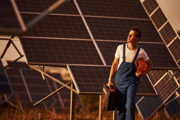 Hält Koffer mit Ausrüstung in der Hand. Männlicher Arbeiter in blauer Uniform im Freien mit Solarbatterien am sonnigen Tag.