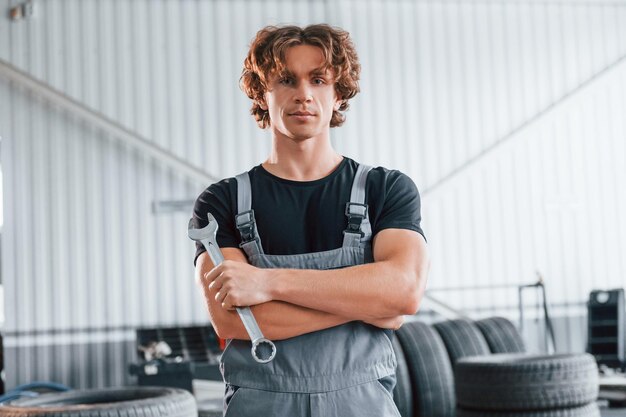 Hält Schraubenschlüssel in der Hand Erwachsener Mann in grauer Uniform arbeitet im Autosalon